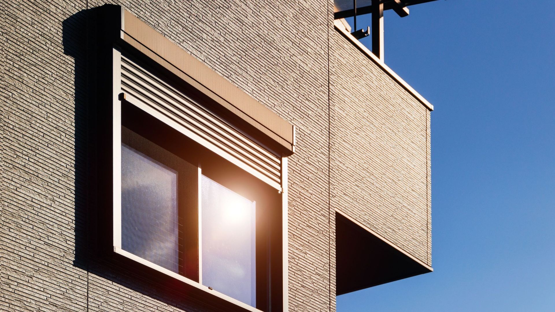 Gebäudefassade mit Fenster und teilweise heruntergelassenem Rollladen unter blauem Himmel im Sonnenlicht