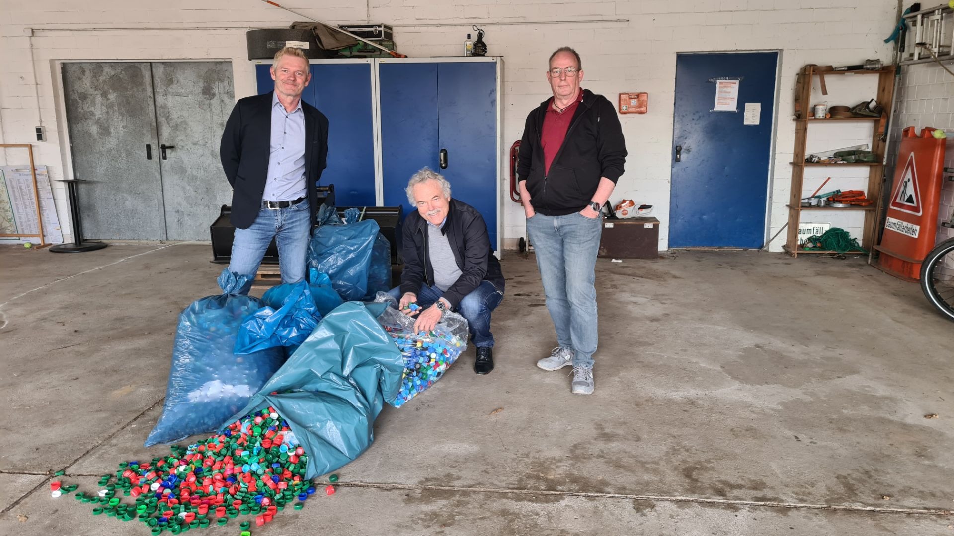 Drei Männer, einer kniend, präsentieren blaue Säcke und bunte Plastikflaschendeckel auf einem Lagerraumboden