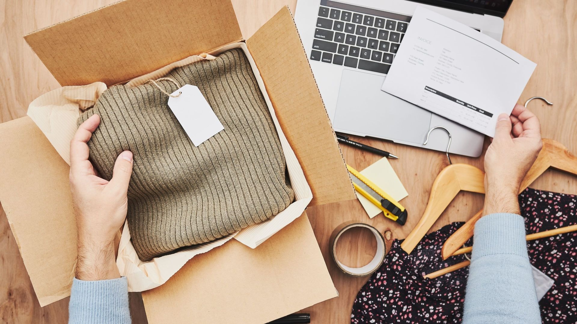 Hände entnehmen einen Pullover aus einem Versandkarton, daneben liegen Rechnung, Laptop und weitere Kleidung