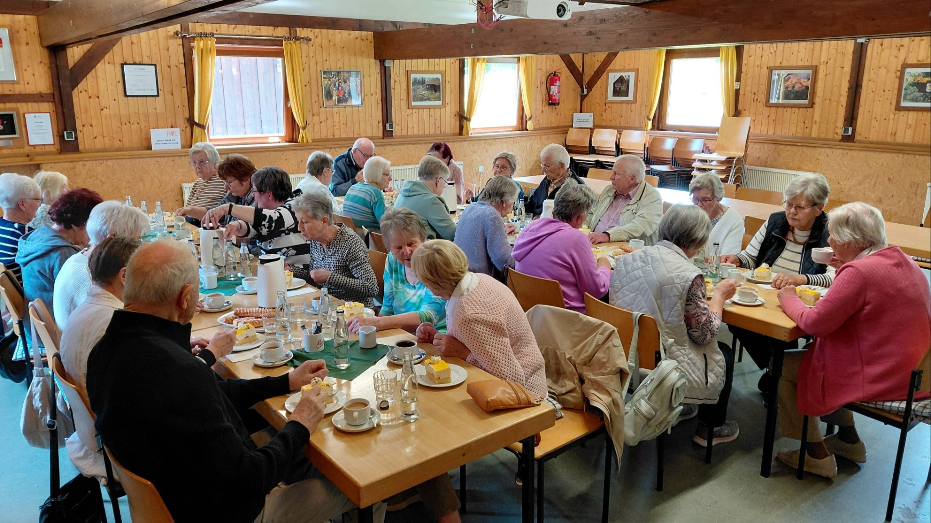 Große Gruppe Senioren sitzt an Holztischen und genießt Kaffee und Kuchen in einem Holzsaal