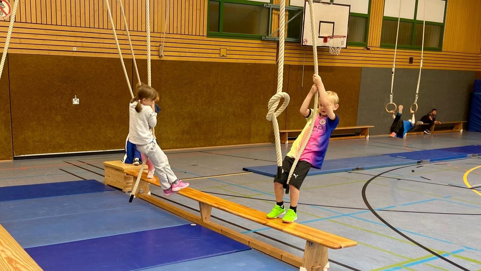 Zwei Kinder balancieren auf einem schmalen Holzbrett und halten sich an herabhängenden Seilen in einer Turnhalle fest
