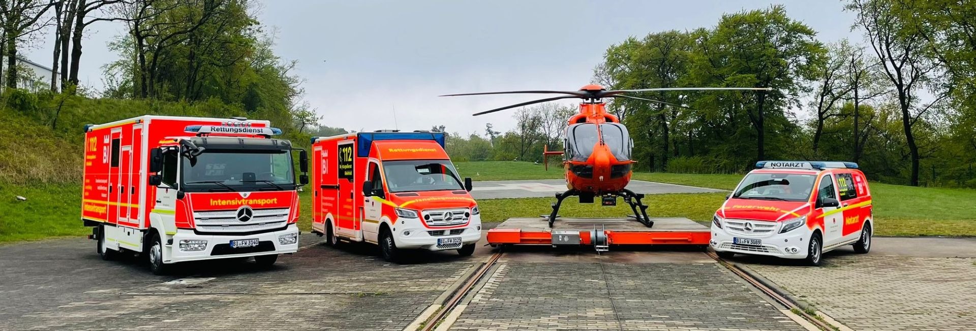 Ein orangefarbener Rettungshubschrauber, ein Intensivtransport-LKW, ein Rettungswagen und ein Notarztfahrzeug auf einer