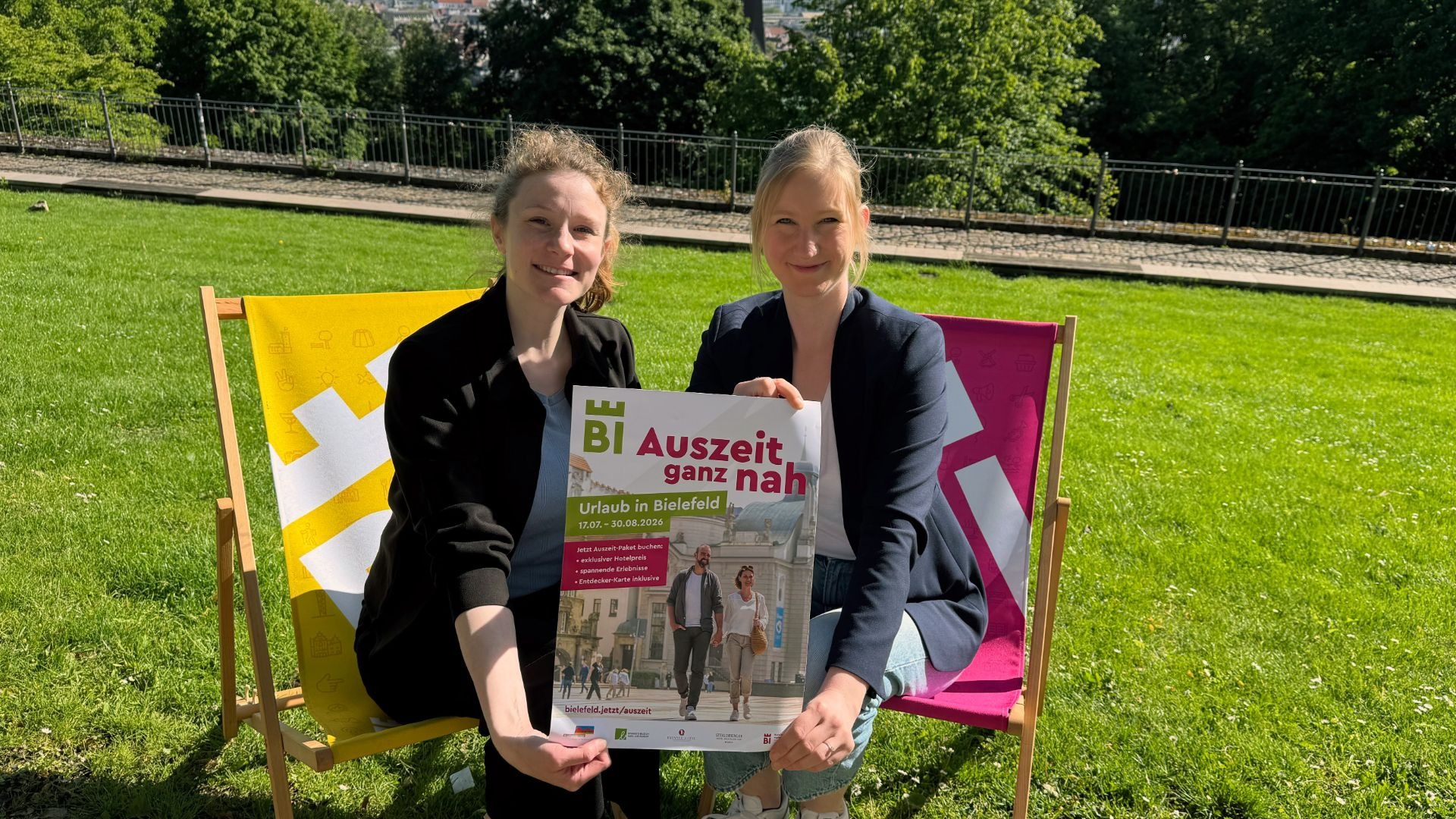 Zwei lächelnde Frauen halten ein Poster für "BI Auszeit ganz nah - Urlaub in Bielefeld" auf einer Wiese