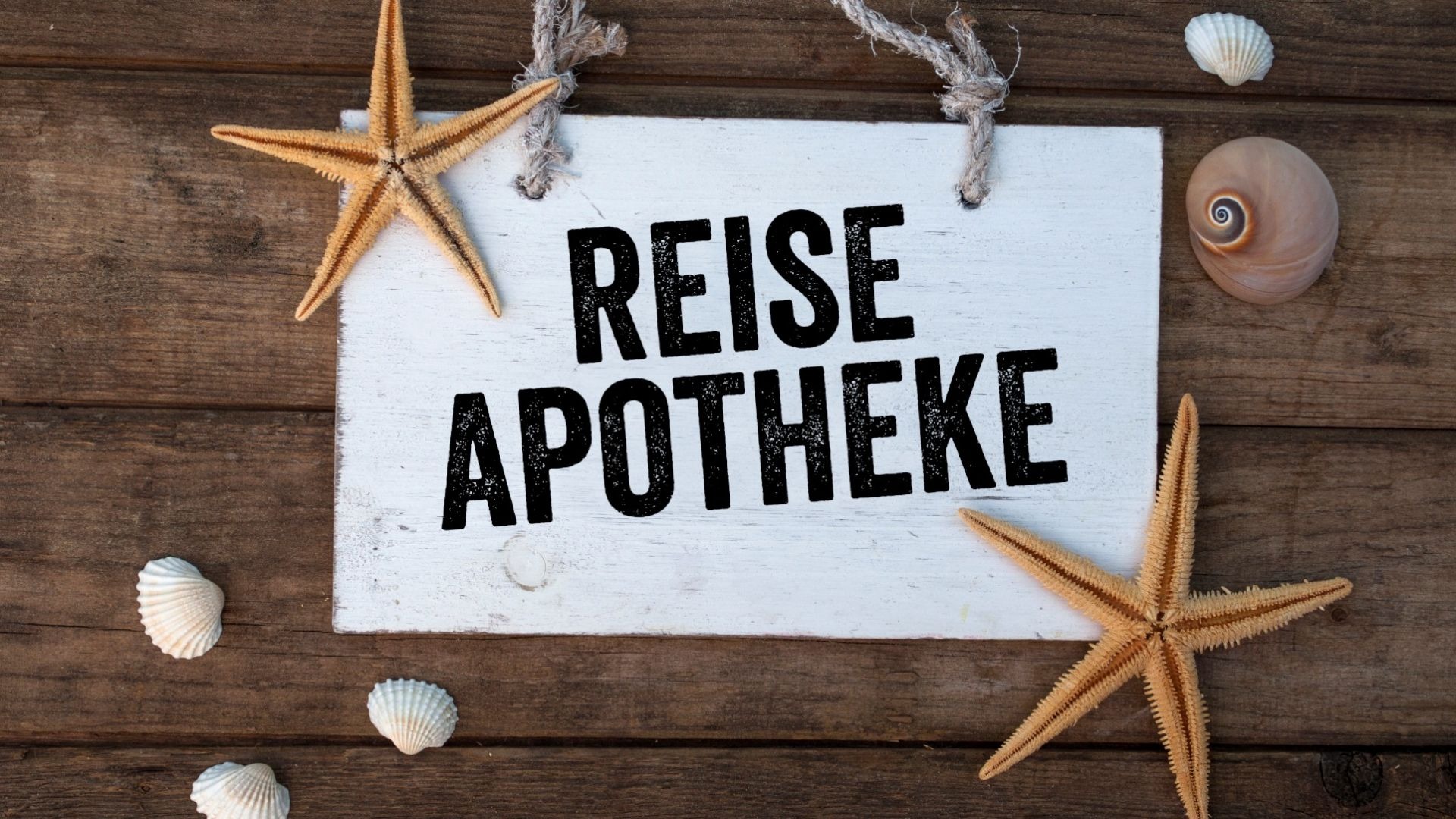 Weißes Holzschild mit der Aufschrift "REISE APOTHEKE" auf braunem Holzuntergrund, umgeben von Seesternen und Muscheln