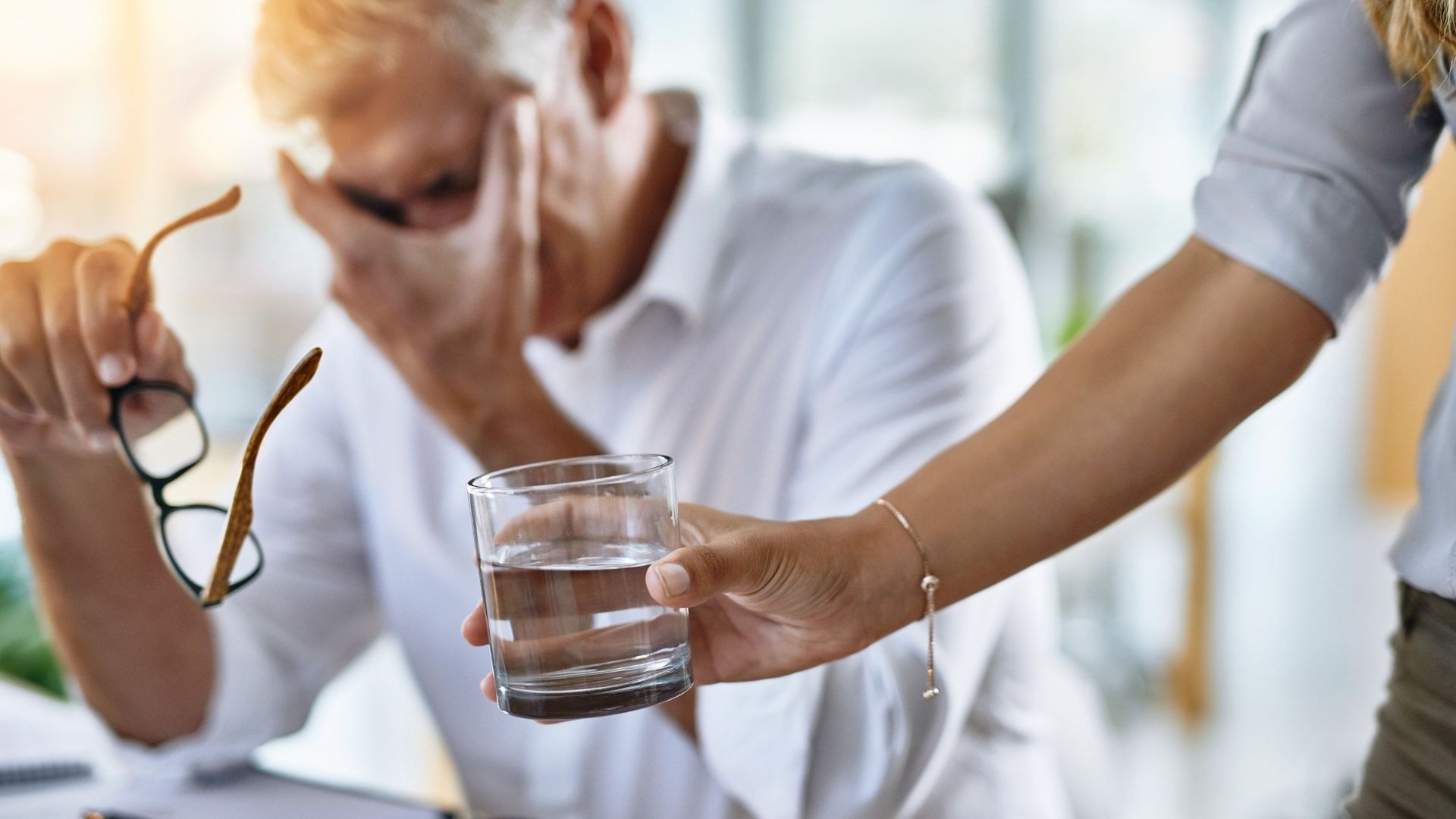 Eine Hand reicht einem Mann, der sich erschöpft an den Kopf fasst, ein Glas Wasser