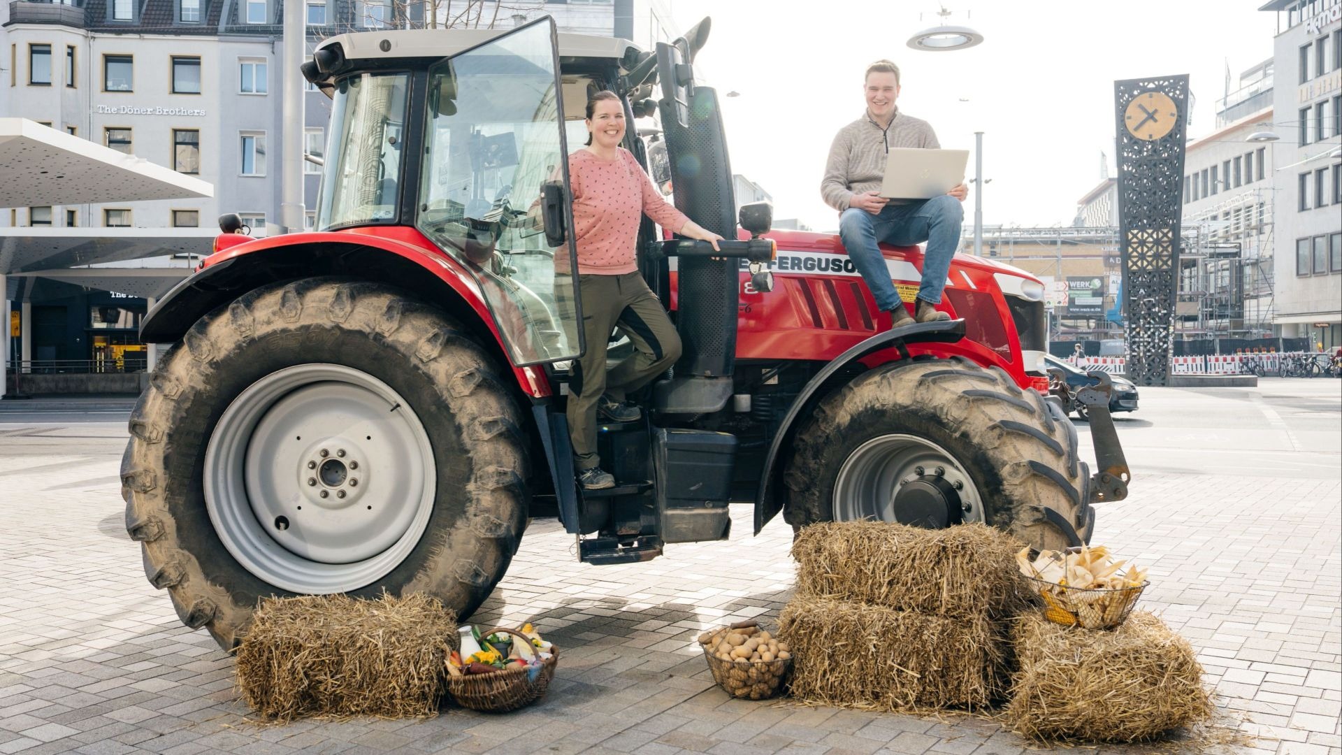 Roter Traktor mit lachender Frau am Einstieg und Mann mit Laptop oben, dazu Heuballen und Körbe mit Gemüse auf