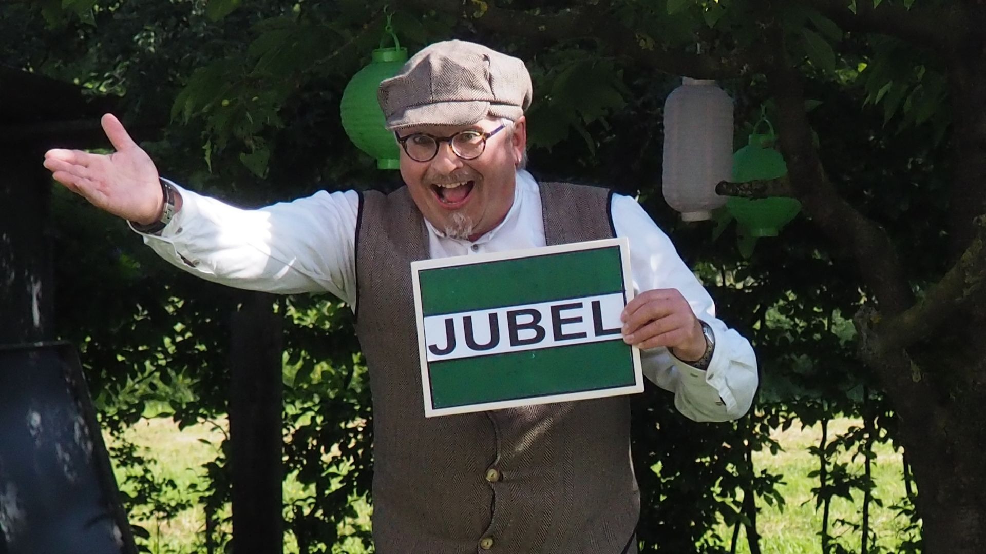 Ein Mann mit Schiebermütze und Weste lacht und hält ein grünes Schild mit der Aufschrift „JUBEL“ unter Bäumen mit