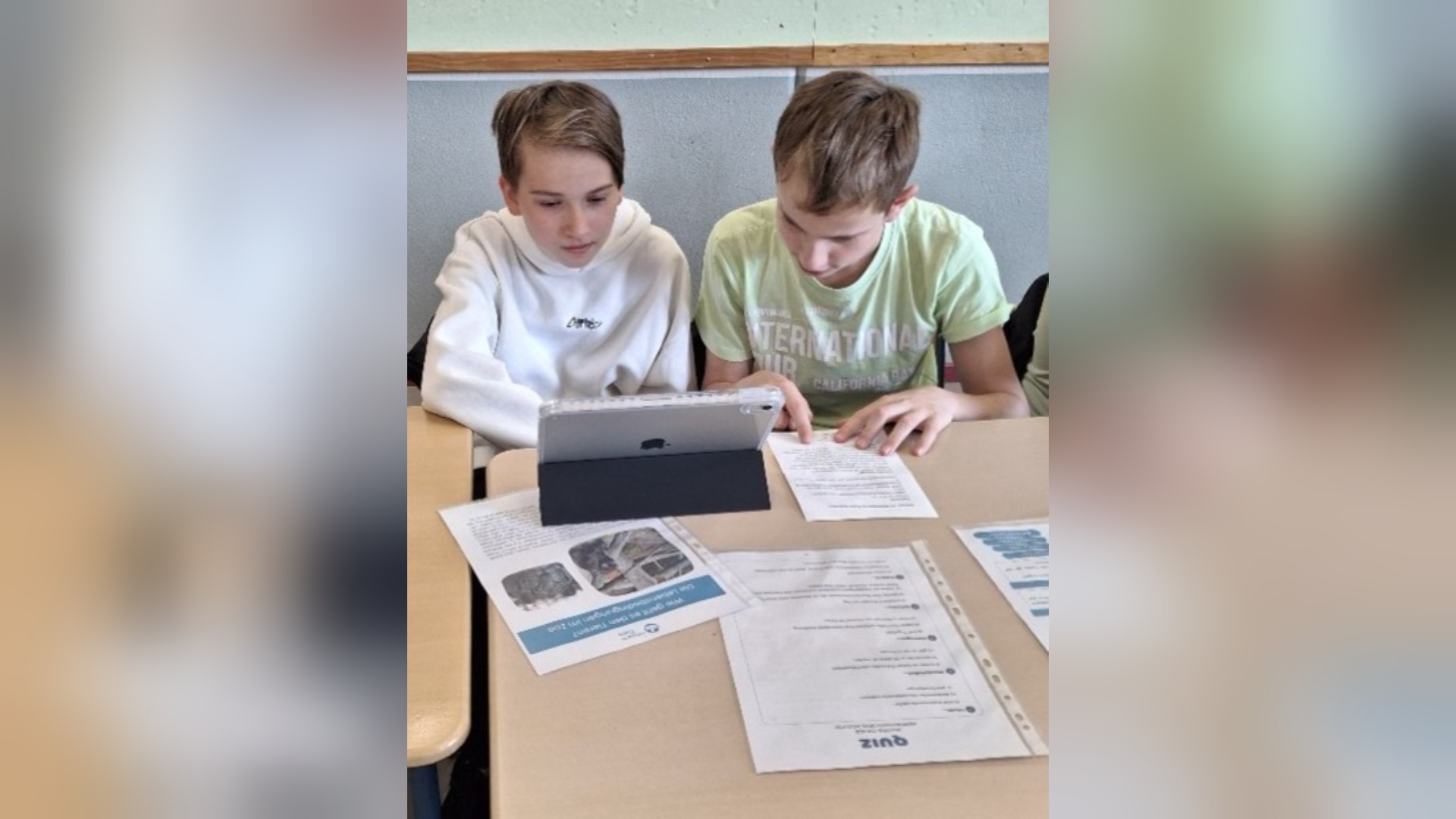 Zwei Schüler sitzen an einem Tisch, betrachten ein Tablet und lesen Arbeitsblätter zum Thema Tierwohl