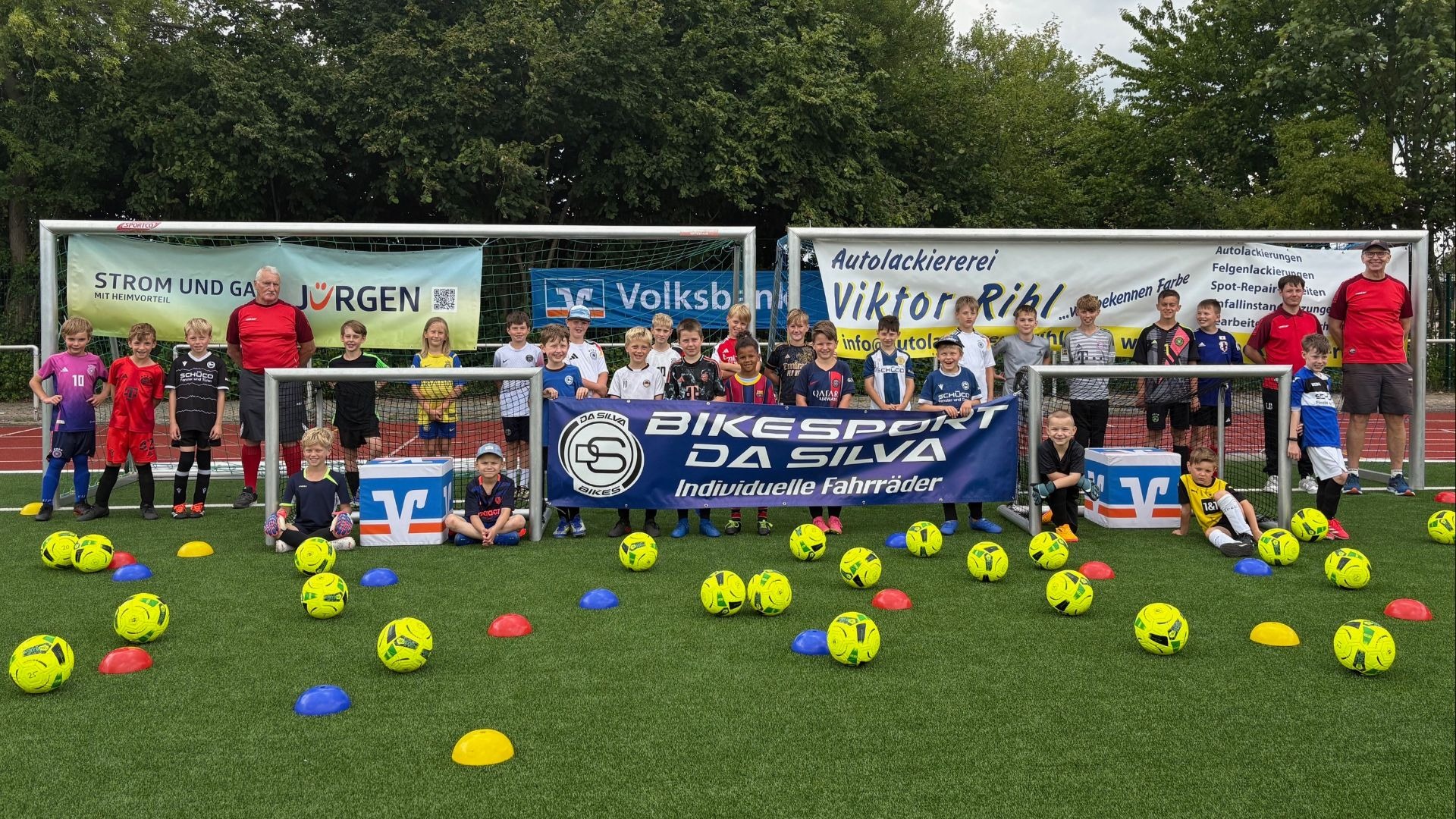 Viele Kinder und einige Erwachsene posieren auf einem Fußballfeld mit Bällen, Toren und Sponsoren-Bannern