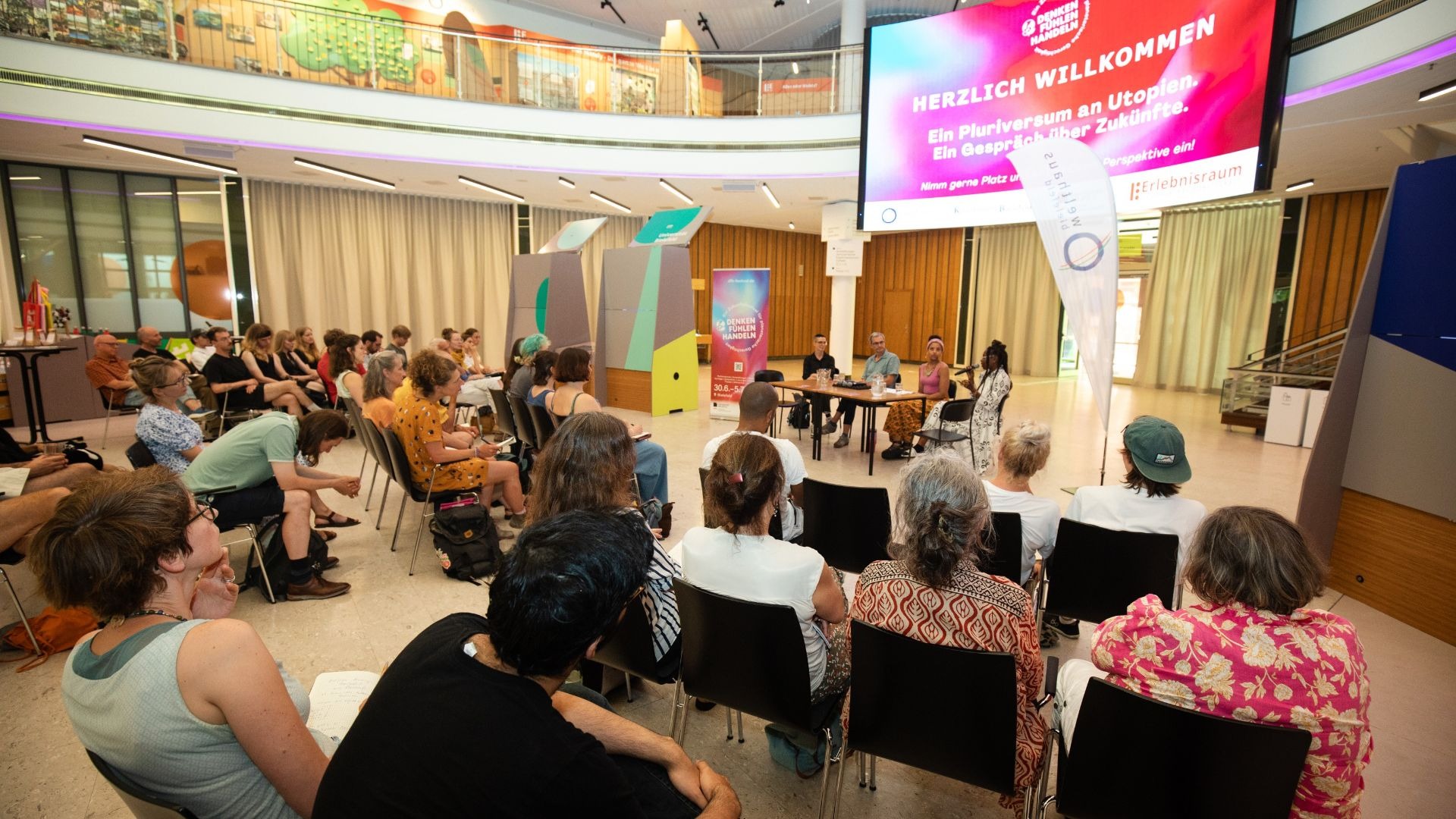 Publikum lauscht Podiumsdiskussion des Festivals „Denken Fühlen Handeln“ vor großem „Herzlich Willkommen“-Bildschirm