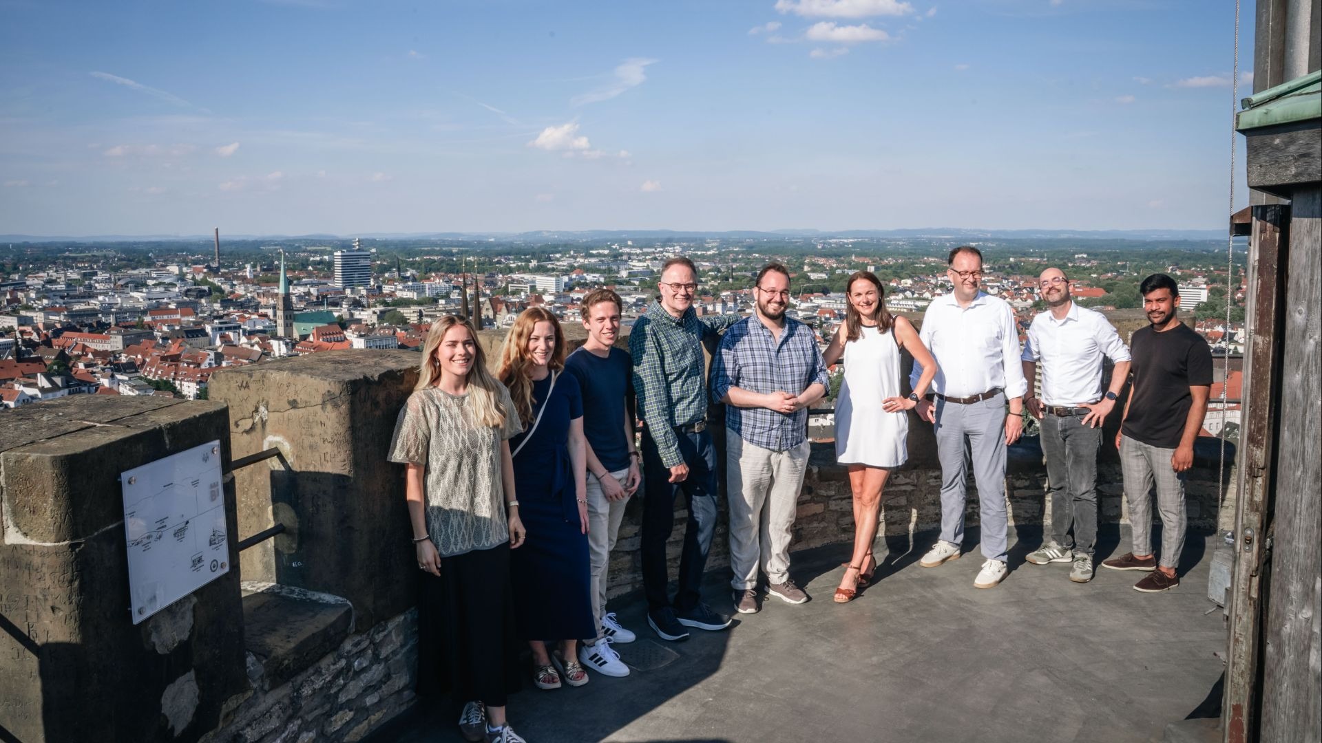 Zehn Menschen stehen lächelnd auf der Sparrenburg mit Panoramablick über die Stadt Bielefeld unter blauem Himmel
