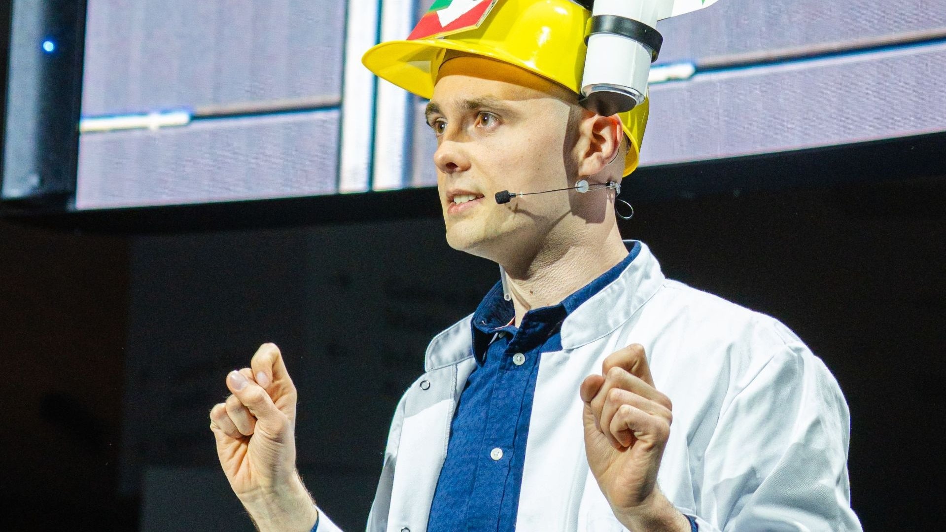 Mann in Laborkittel und auffälligem gelben Helm mit Mikrofon spricht und gestikuliert auf Bühne