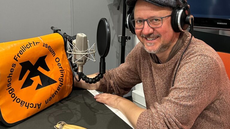 Lächelnder Mann mit Kopfhörern und Mikrofon in einem Aufnahmestudio neben einer Tasche des Museums Oerlinghausen