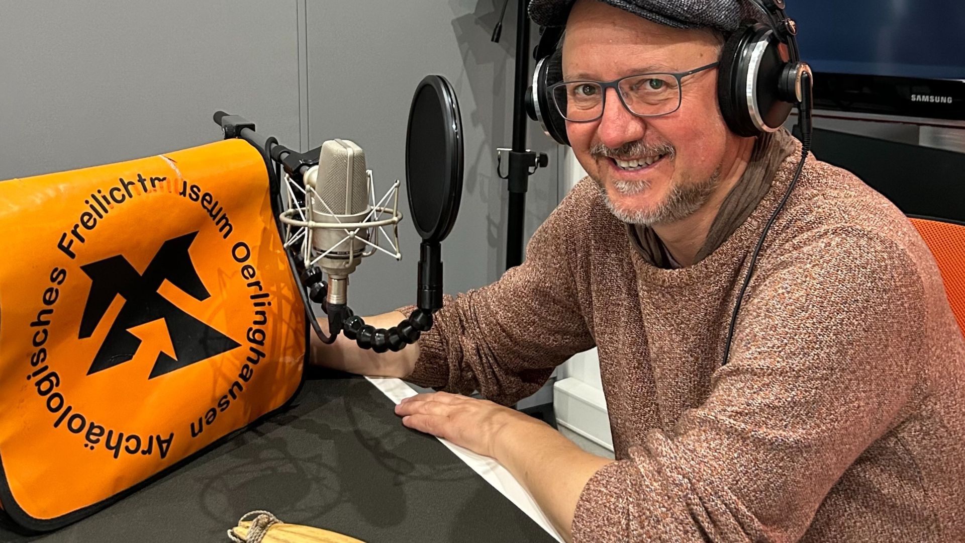 Lächelnder Mann mit Kopfhörern und Mikrofon in einem Aufnahmestudio neben einer Tasche des Museums Oerlinghausen