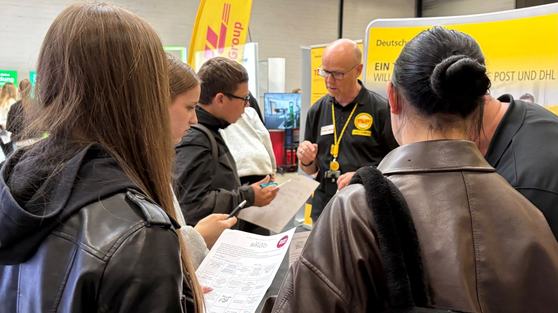 Schüler an einem Berufsmesse-Stand bei Deutsche Post und DHL, die einem Mitarbeiter zuhören und Informationsblätter