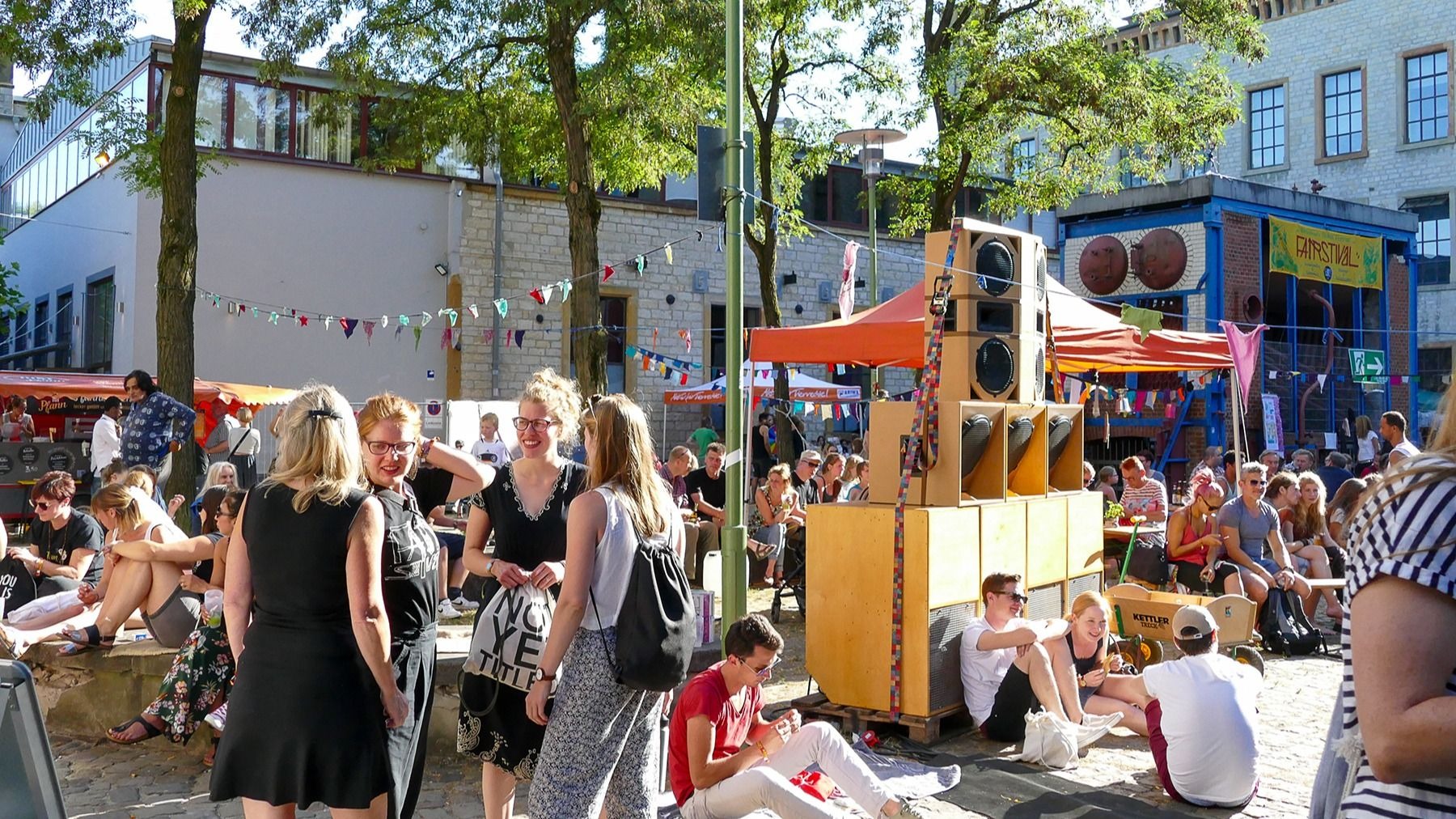 Menschen bei einem Open-Air-Festival mit großen Lautsprechern, Zelten und einem Fairstival-Banner im Hintergrund