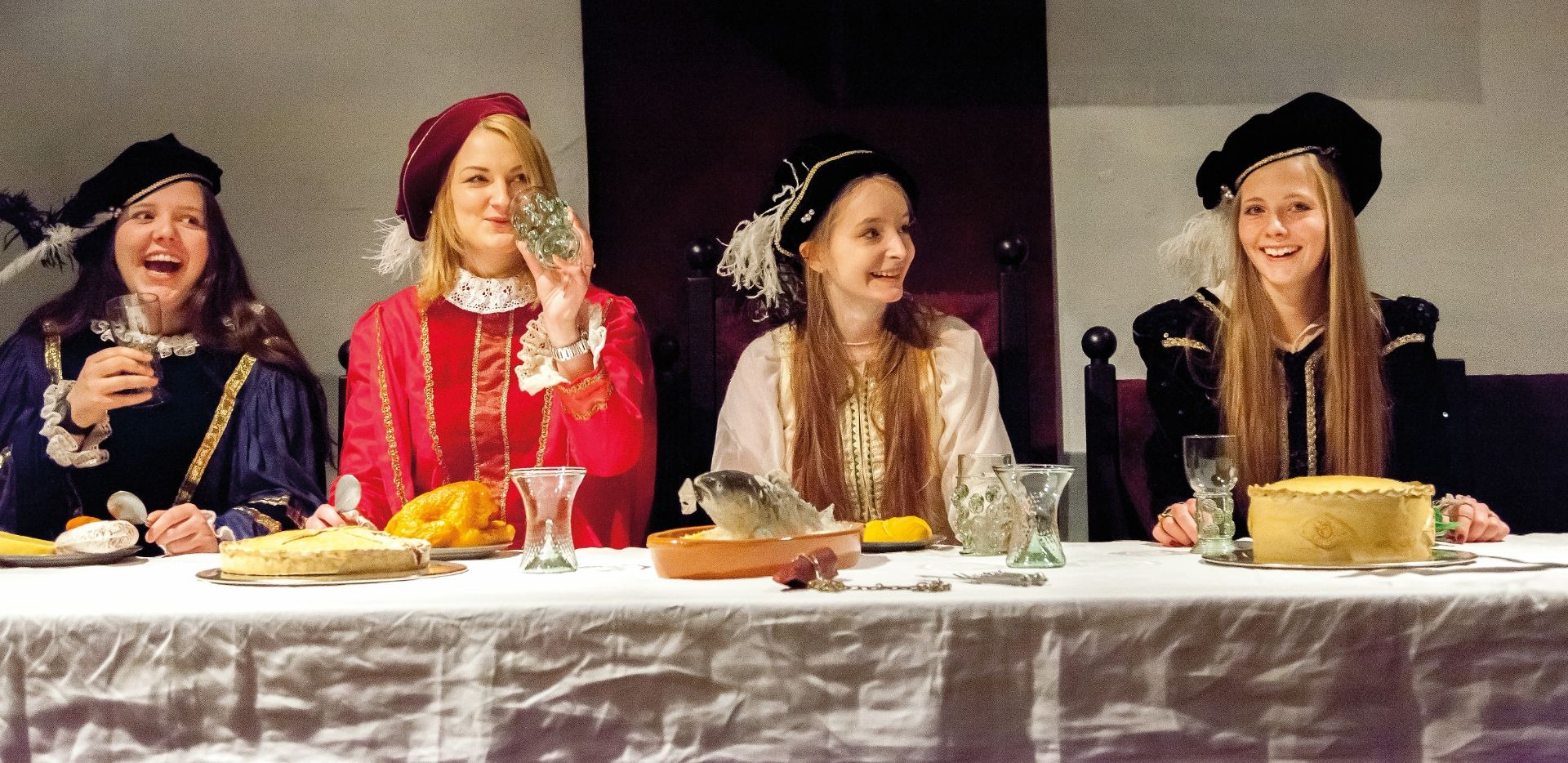 Vier Personen in historischen Renaissance-Gewändern speisen lachend an einem festlich gedeckten Tisch mit Gebäck und