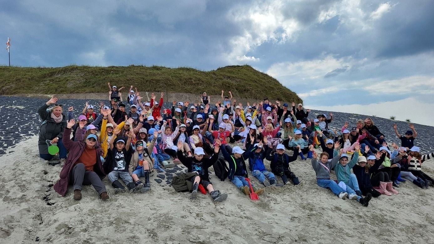 Viele Kinder und mehrere Erwachsene sitzen auf einem Sandhang am Strand und winken vor einer grasbewachsenen Düne