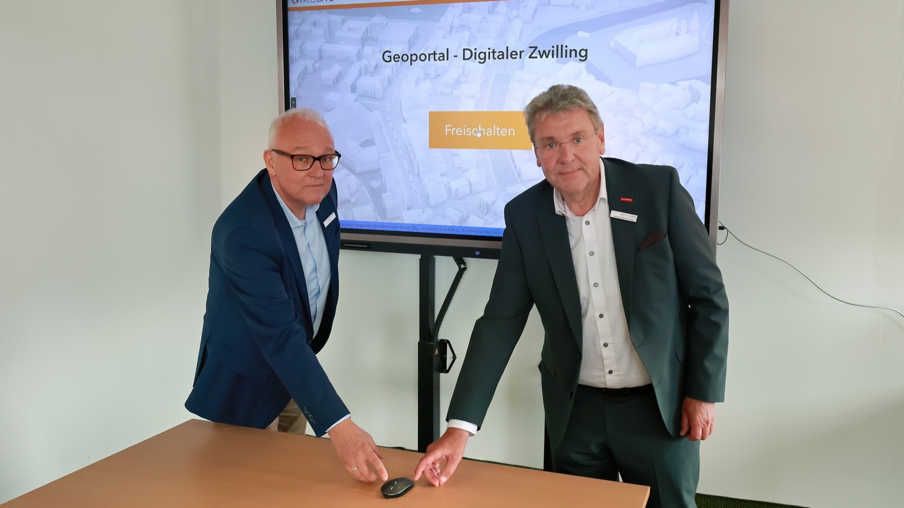 Zwei Männer in Anzügen zeigen auf Computermaus vor Bildschirm mit 'Geoportal - Digitaler Zwilling' und 'Kreis Lippe