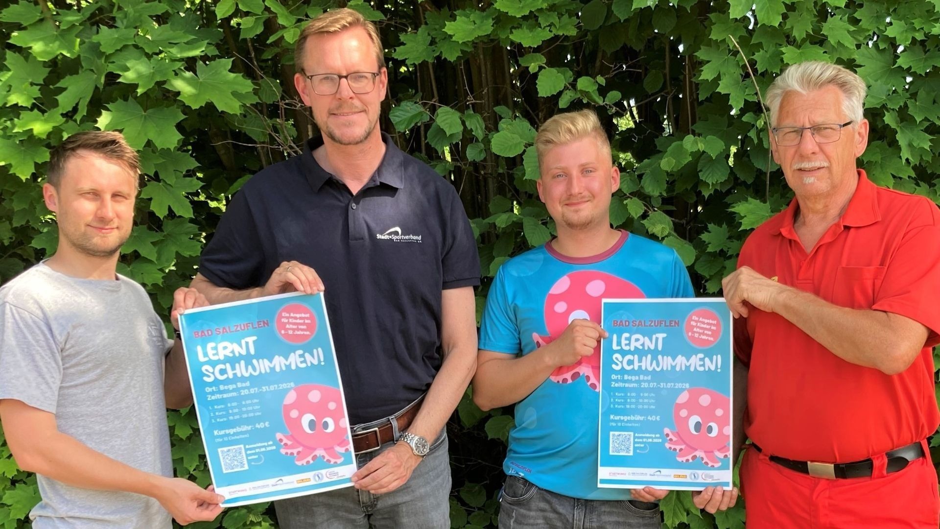 Vier Männer stehen vor grünen Büschen und halten zwei Poster für den Schwimmkurs "Bad Salzuflen lernt Schwimmen