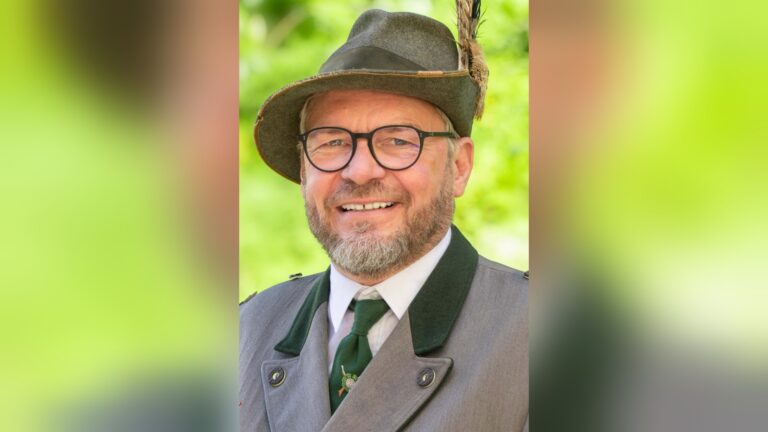 Lachender Mann mit Bart, Brille, grauem Trachtenanzug, Trachtenhut mit Feder, vor grünem Hintergrund