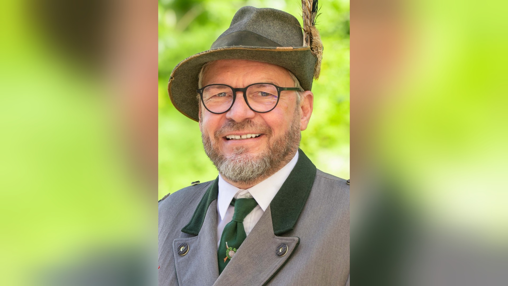 Lachender Mann mit Bart, Brille, grauem Trachtenanzug, Trachtenhut mit Feder, vor grünem Hintergrund