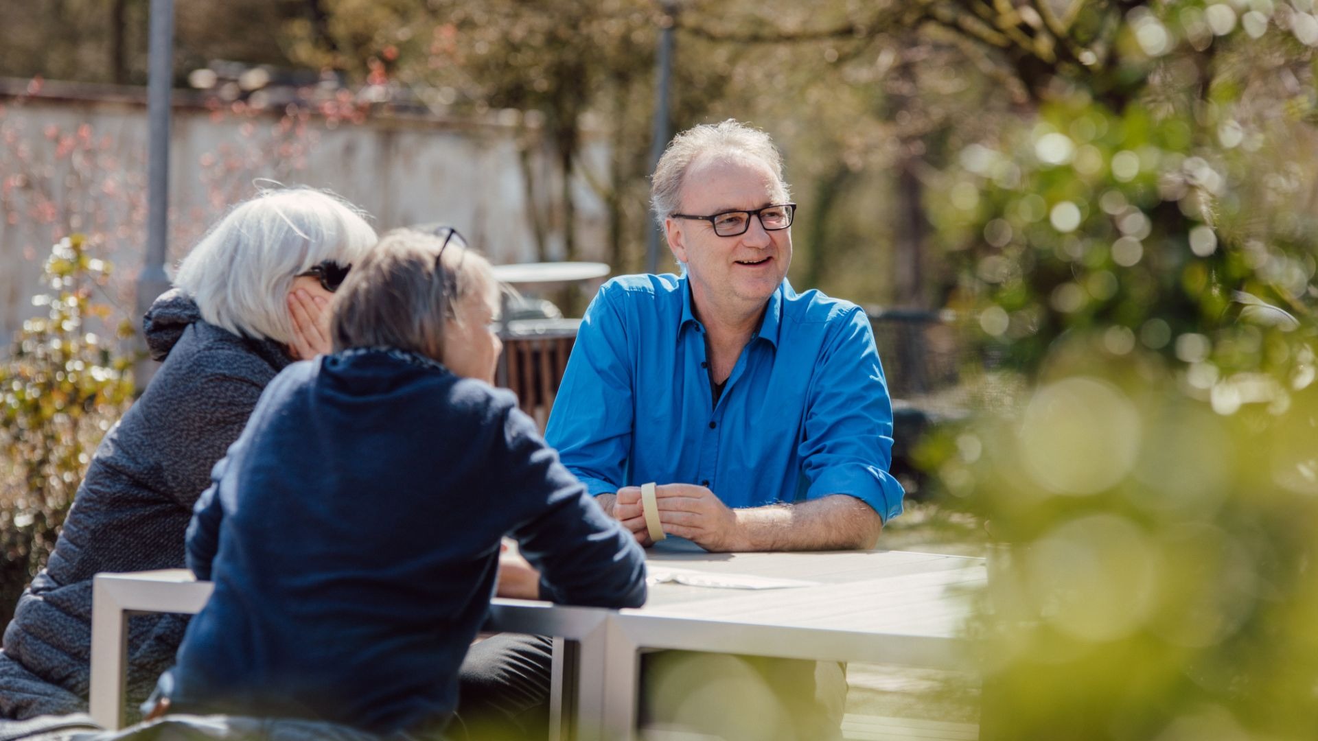 Drei Personen sitzen an einem Tisch im Freien, ein Mann in blauem Hemd lacht und unterhält sich mit zwei anderen