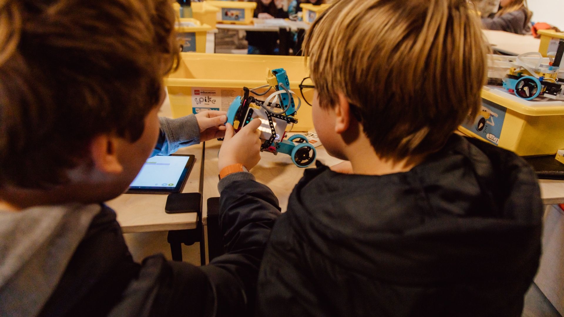 Zwei Kinder arbeiten an einem Tisch: Eines baut einen blauen und gelben Roboter, das andere bedient ein Tablet