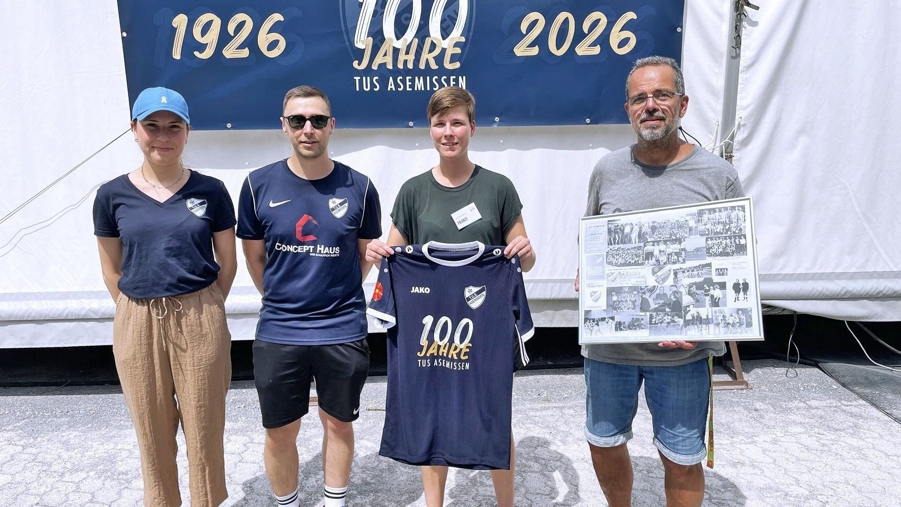 Vier Personen stehen vor einem "100 Jahre TUS ASEMISSEN" Banner, eine hält ein Jubiläumstrikot, eine andere eine