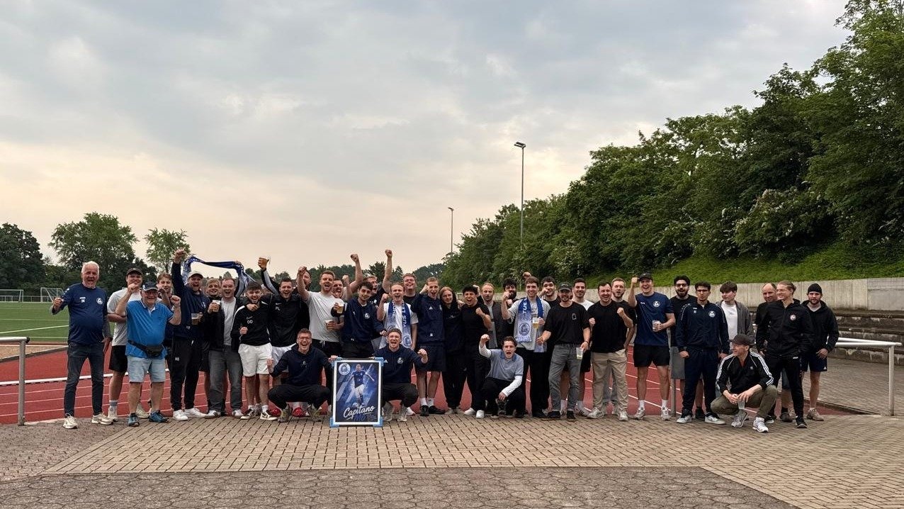 Große Gruppe jubelnder Fußballer mit erhobenen Fäusten, Getränken und einem Poster am Fußballfeld