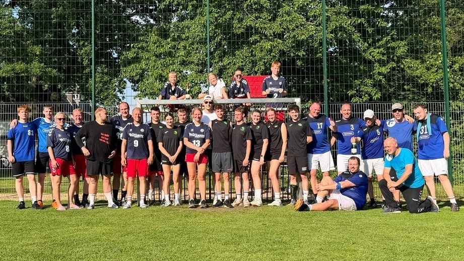 Lachende Gruppe Handballer und Handballerinnen posiert mit Pokal auf Sportplatz vor Tor