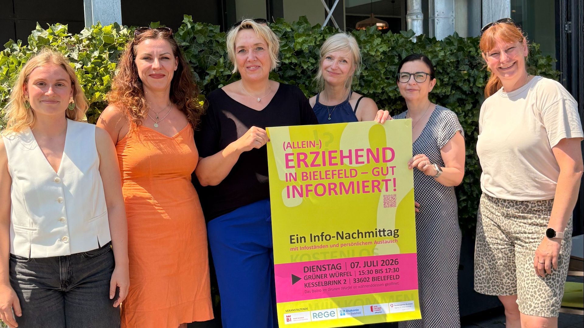 Sechs Frauen lächeln vor Efeuwand, zwei halten gelbes Plakat für Info-Nachmittag für (Allein-)Erziehende in Bielefeld