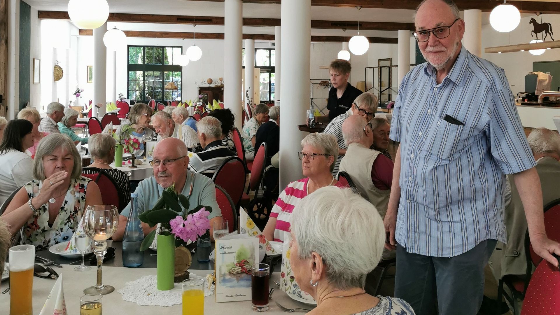 Eine Gruppe Senioren sitzt an Tischen in einem Restaurant, ein Mann mit Brille und Hemd steht im Gang