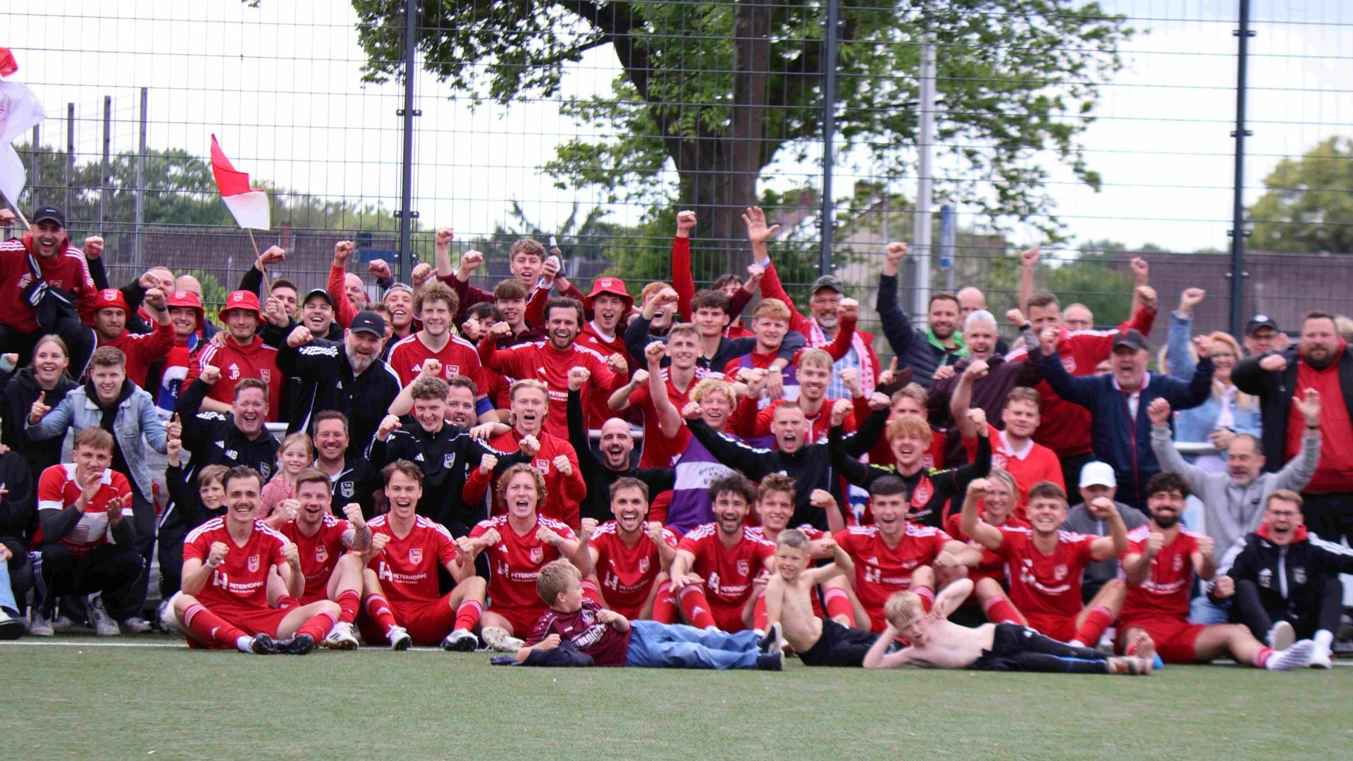 Große Gruppe feiernder Fußballspieler und Fans in roten Trikots auf einem Kunstrasenplatz
