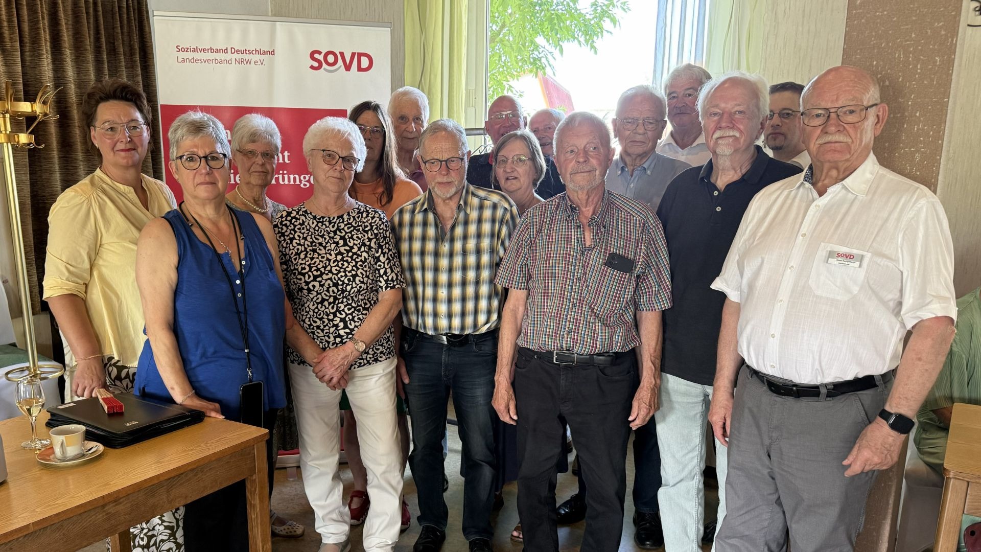 Eine Gruppe älterer Frauen und Männer des SoVD stehen in einem Raum mit einem SoVD-Banner im Hintergrund