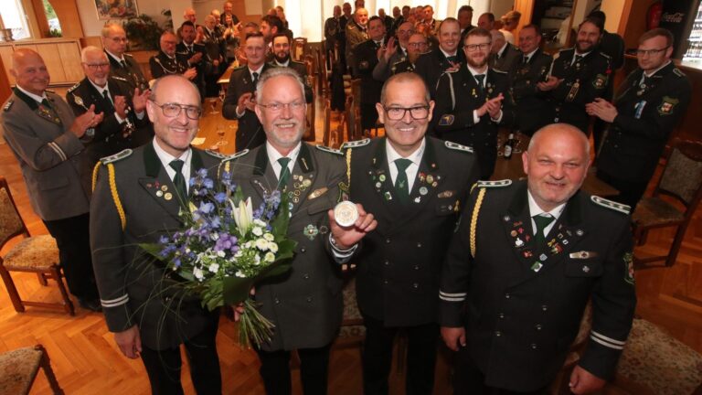 Vier Männer in dunklen Uniformen lächeln mit Blumenstrauß und Medaille, andere applaudieren im Saal