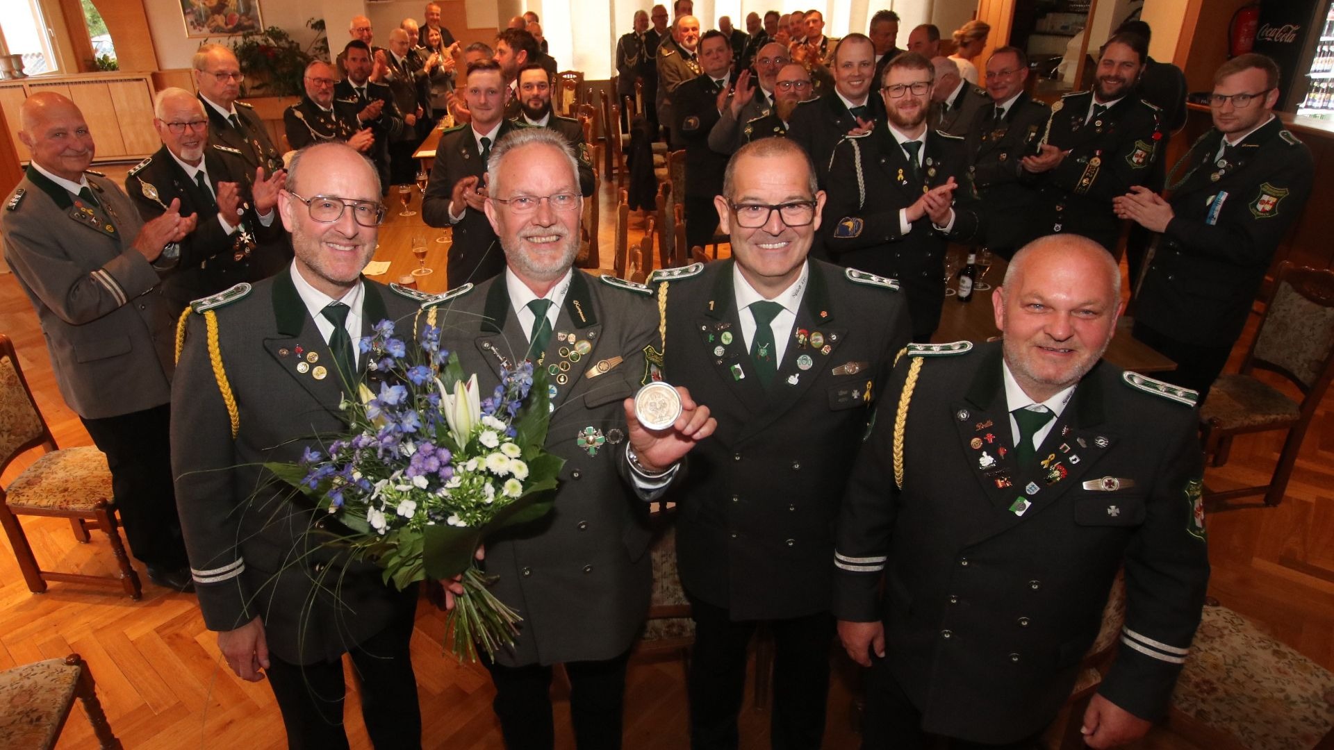 Vier Männer in dunklen Uniformen lächeln mit Blumenstrauß und Medaille, andere applaudieren im Saal