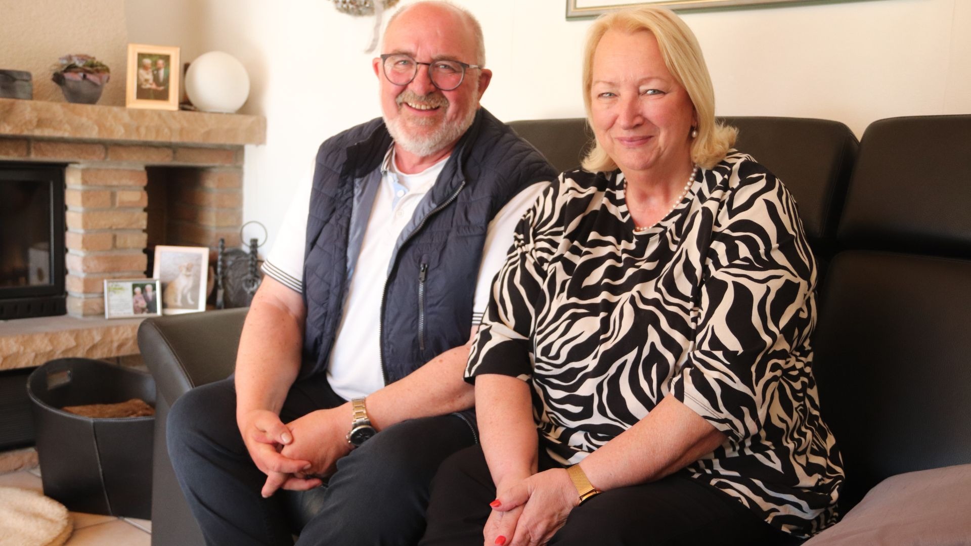 Marion und Bernd Oberschelp sitzen lächelnd auf einem dunklen Sofa, halten Hände, im Hintergrund ein Kamin