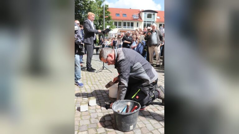 Mann verlegt Stolpersteine auf gepflastertem Platz, während ein Redner spricht und Menschen zuschauen