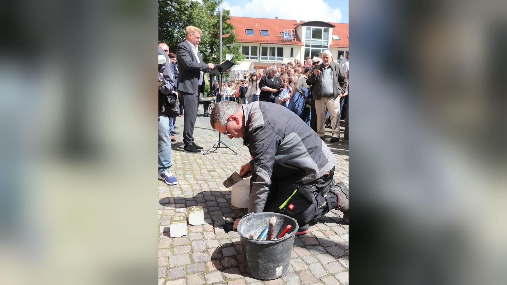 Mann verlegt Stolpersteine auf gepflastertem Platz, während ein Redner spricht und Menschen zuschauen