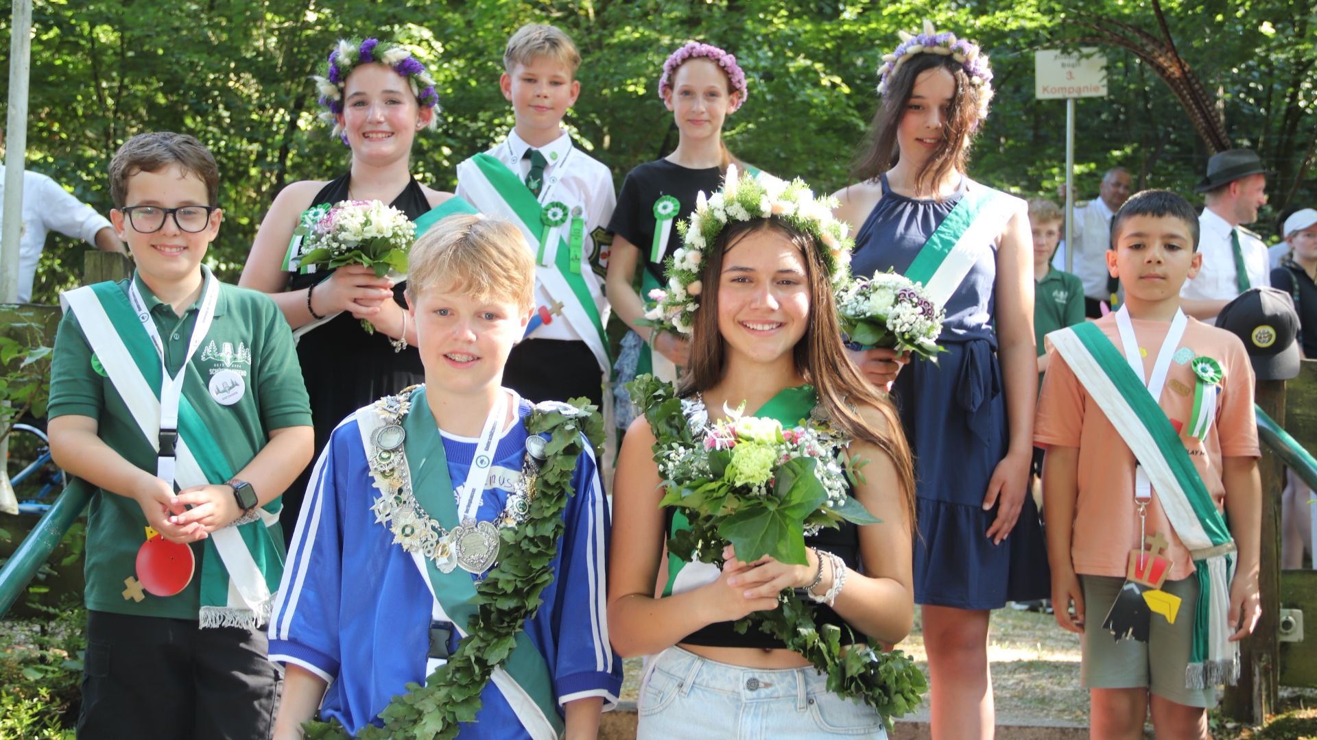 Eine Gruppe festlich gekleideter lachender Kinder mit grünen Schärpen und Blumenkränzen, der Kinderschützenkönig trägt