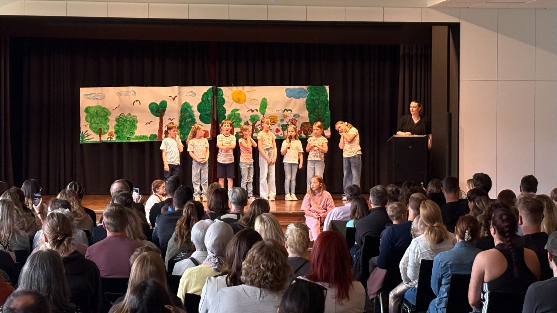 Schüler treten auf einer Bühne vor einem bemalten Bühnenbild und Publikum auf, eine Frau steht am Rednerpult