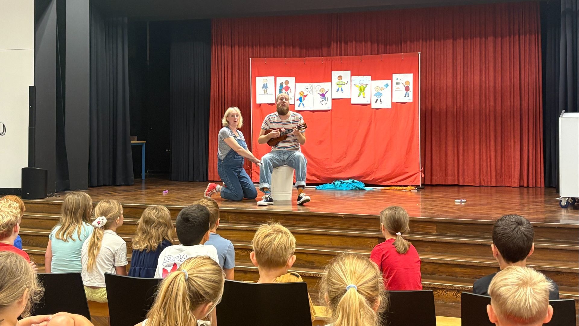 Mann spielt Ukulele und Frau kniet auf Bühne vor Kindern, Hintergrund rote Vorhänge mit vielen Kinderzeichnungen