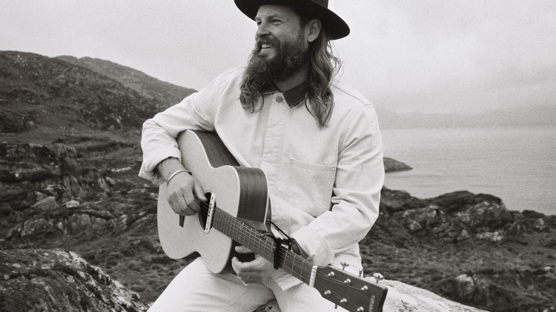 Ian Hooper mit Bart und Hut spielt lächelnd akustische Gitarre in einer felsigen Küstenlandschaft am Meer