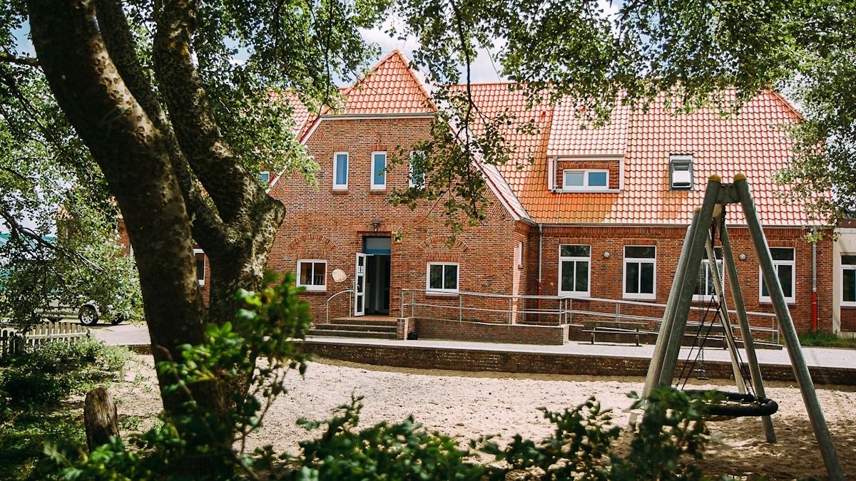 Ziegelsteingebäude des Haus Detmold auf Norderney mit roten Dächern, umgeben von Bäumen und einem Sandspielplatz mit