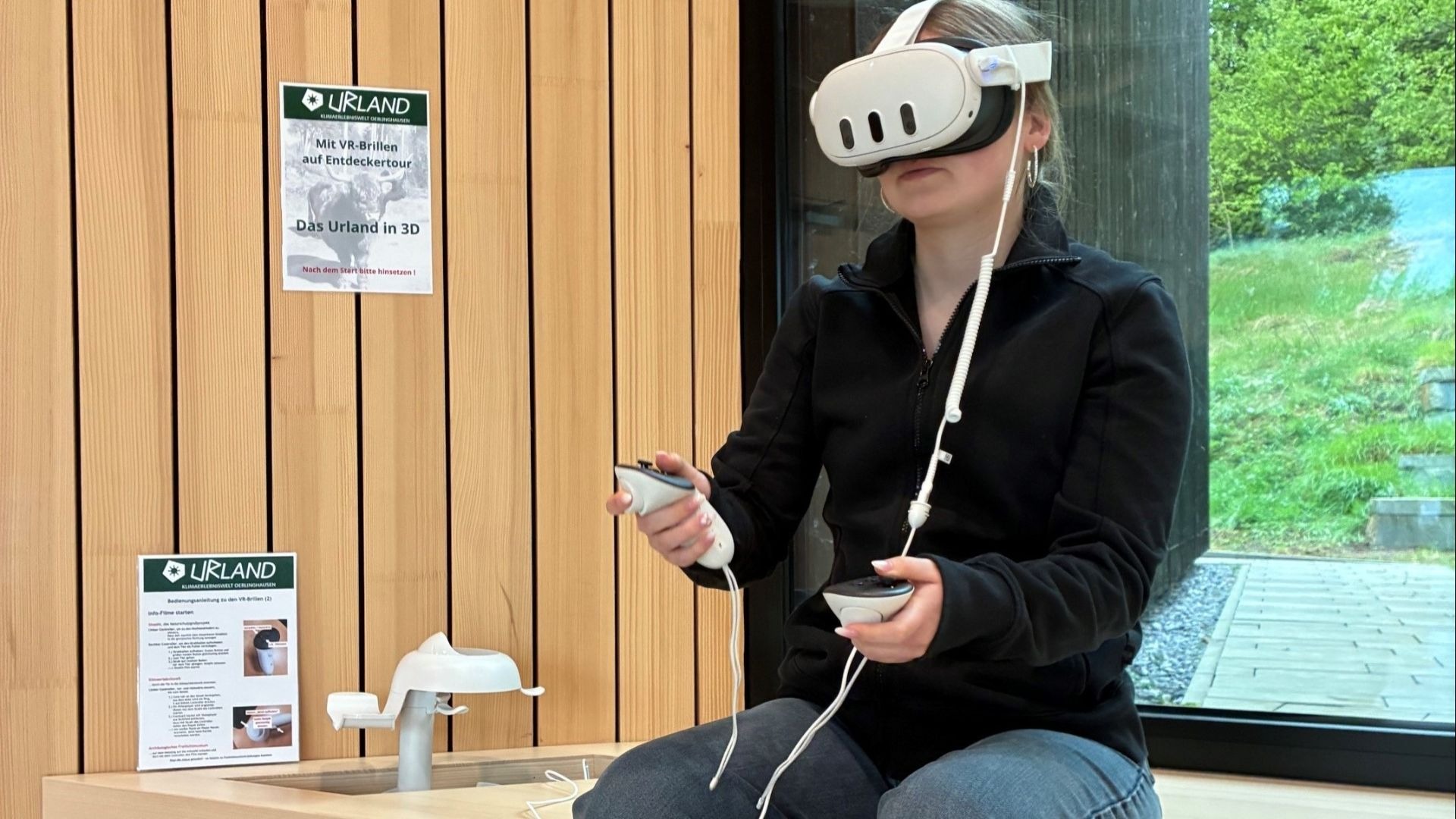 Person trägt VR-Brille und hält Controller in der Klimaerlebniswelt Oerlinghausen, vor Holzwand und Fenster mit Natur