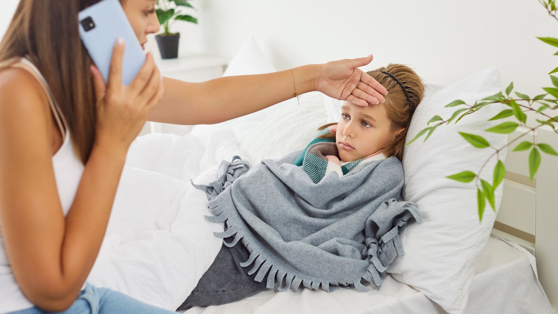 Eine Frau telefoniert und fühlt die Stirn eines kranken, zugedeckten Mädchens im Bett