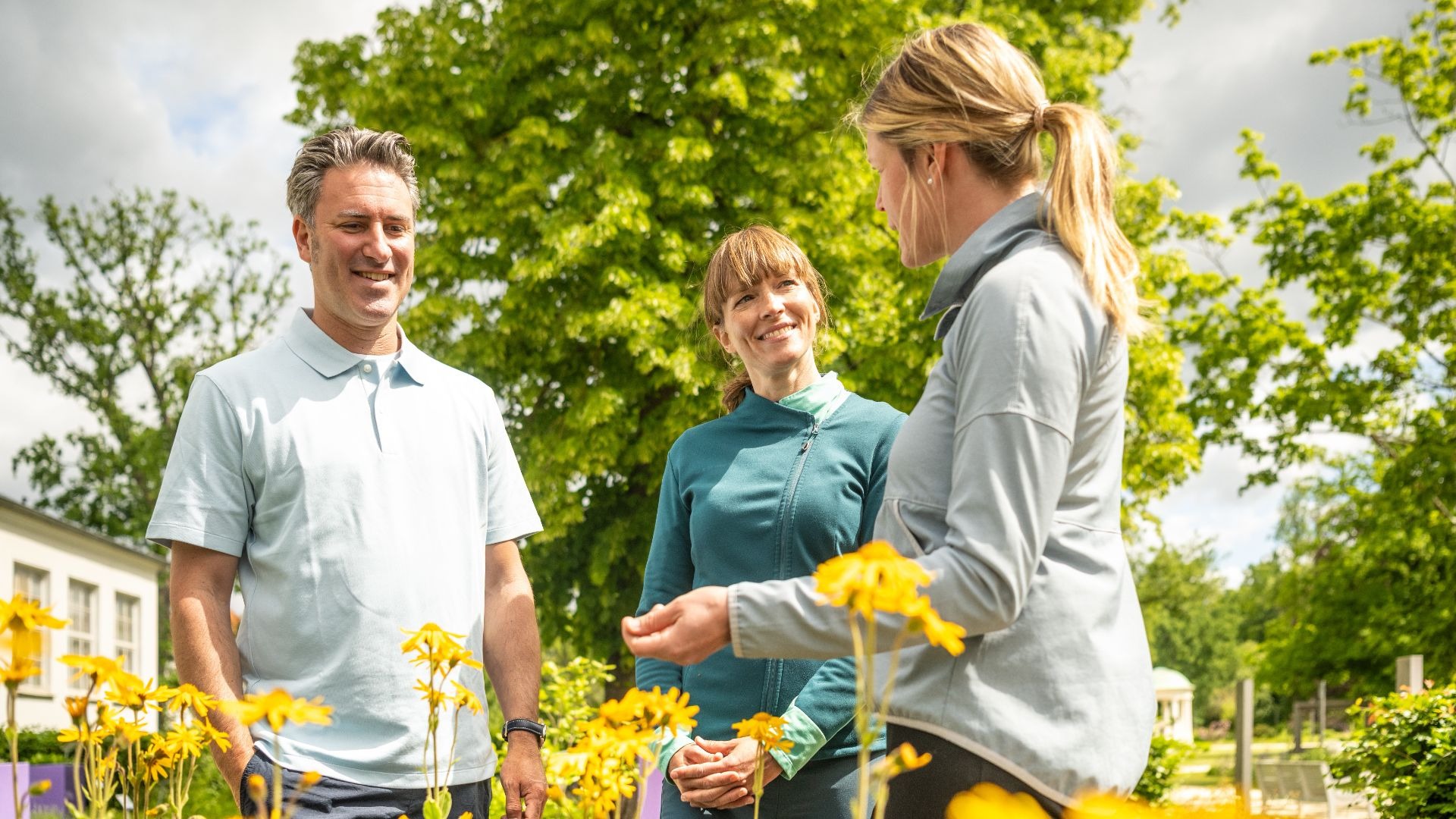 Drei Personen unterhalten sich in einem sonnigen Park mit gelben Blumen und Bäumen