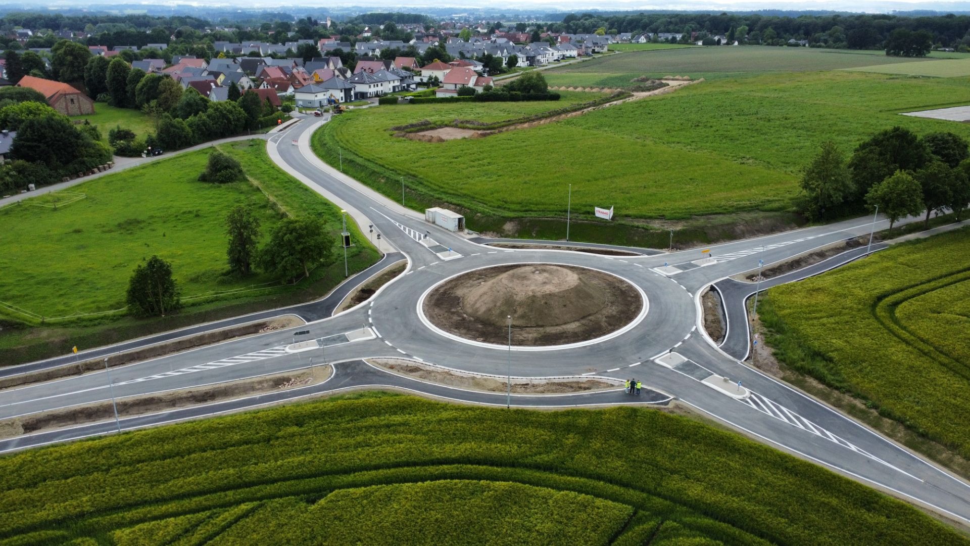 Neue Kreisverkehrsanlage mit Erdhügel, mehreren Zufahrtsstraßen, Feldern, einem Wohngebiet und zwei Arbeitern