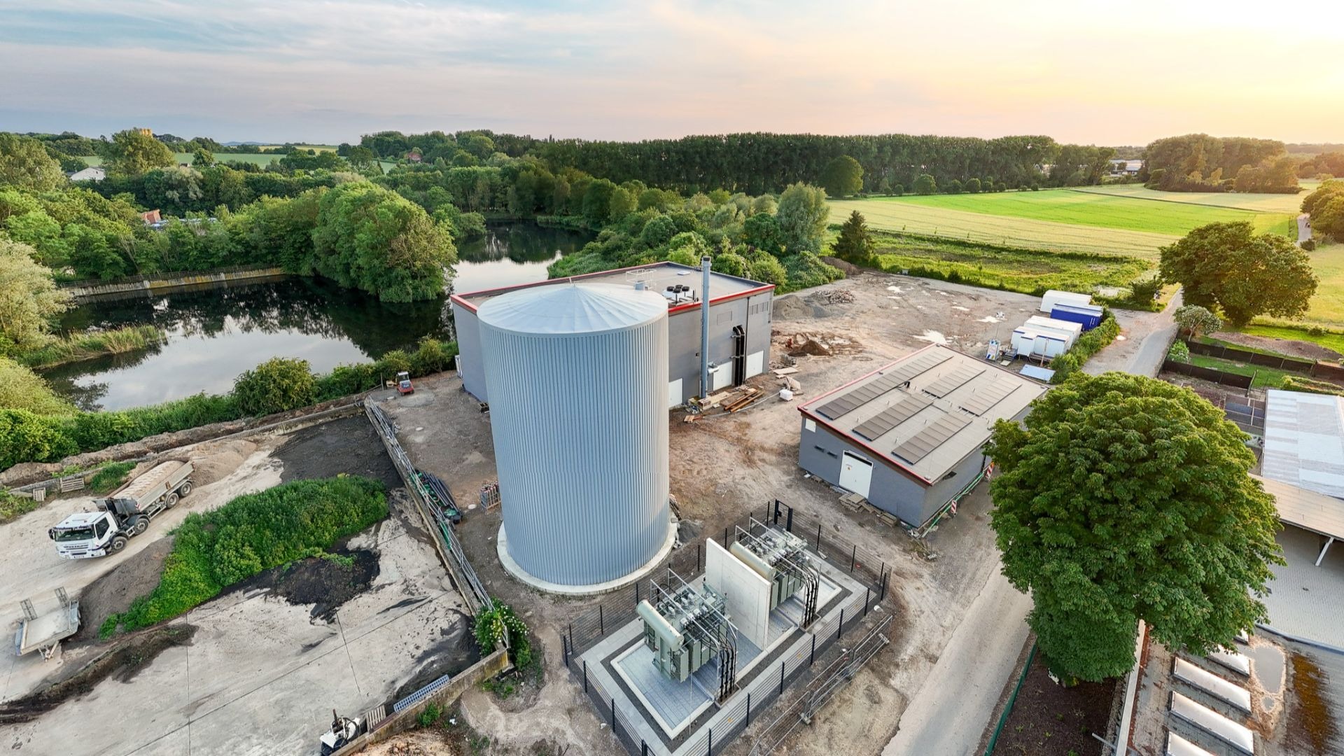 Luftbild einer Energieanlage mit Umspanntechnik, großem Silo, Gebäuden, einem See und grünen Feldern